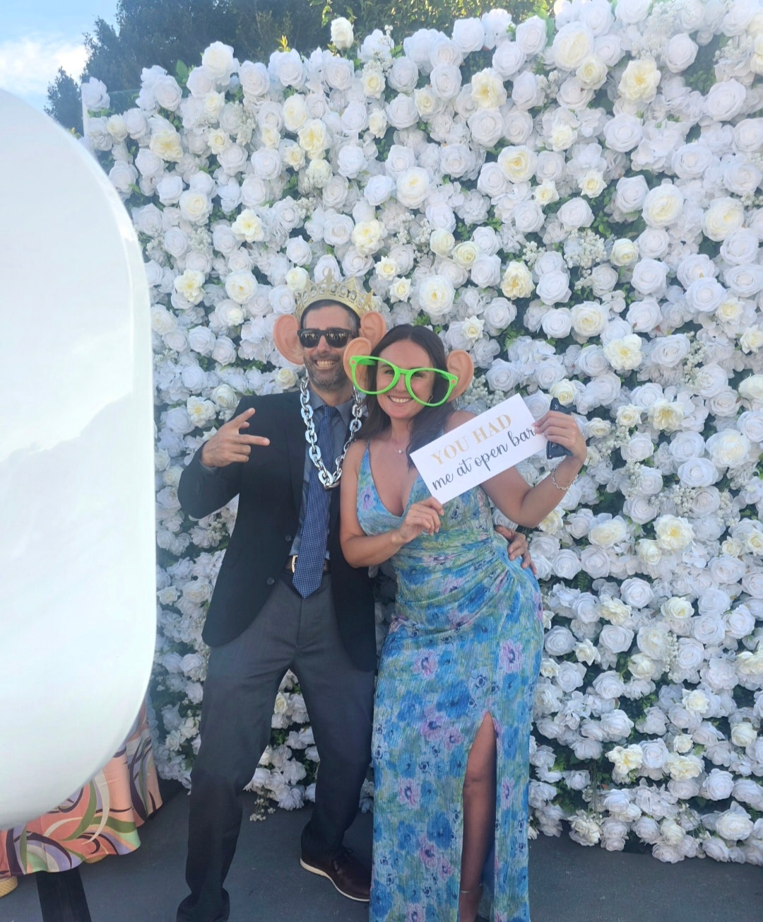 Couple posing with props in front of white flower wall at wedding